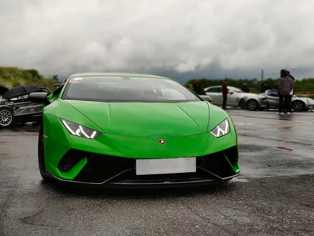 Grøn Lamborghini sportsvogn parkeret på en våd vej med flere biler og mennesker i baggrunden.