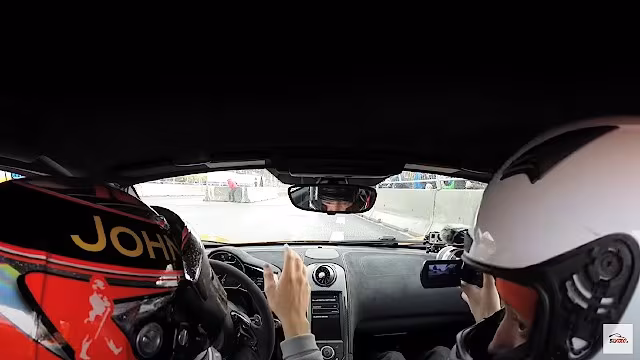 To personer i McLaren 650S racerbil iført hjelme, set fra bilens kabine, mens de er på en bane.