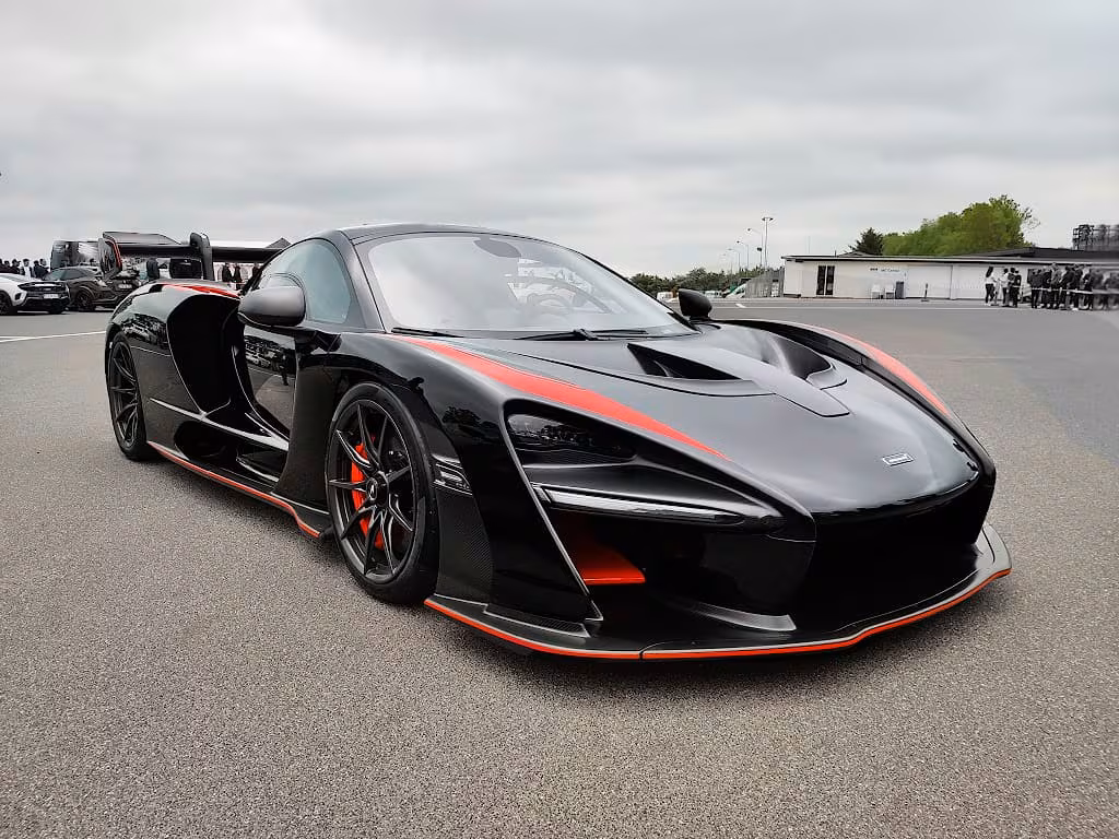 Sort og rød Mclaren Senna sportsvogn på parkeringsplads under overskyet himmel.