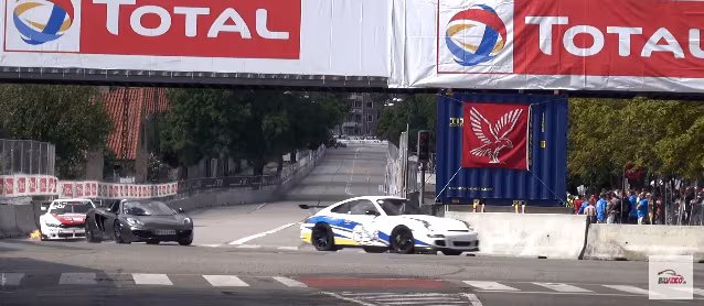 Hvid og blå Porsche 911 racerbil kører gennem et gadesving, mens to andre biler nærmer sig bagfra.